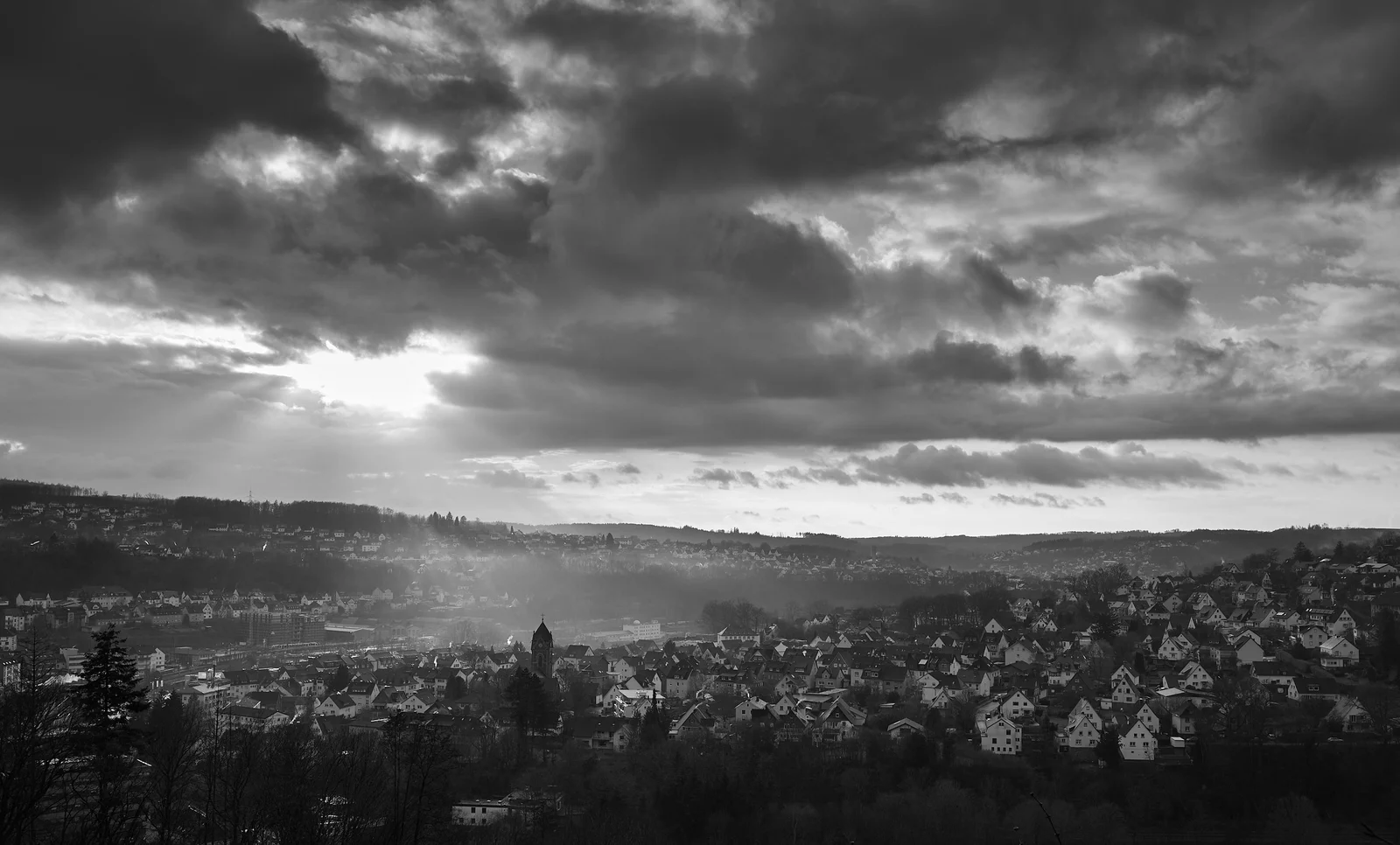 Dunkle Wolken und helle Sonnenstrahlen über einem Ort zwischen bewaldeten Hügeln, in Schwarzweiß.